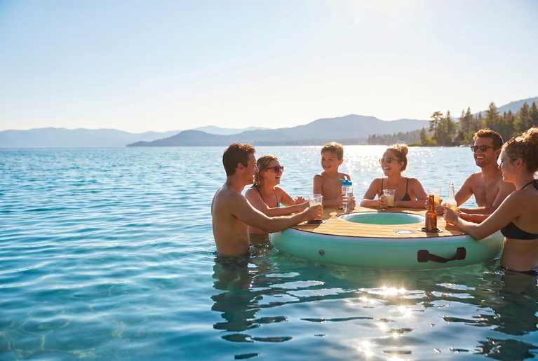People enjoying drinks around a floating bar on a lake with mountains in the background